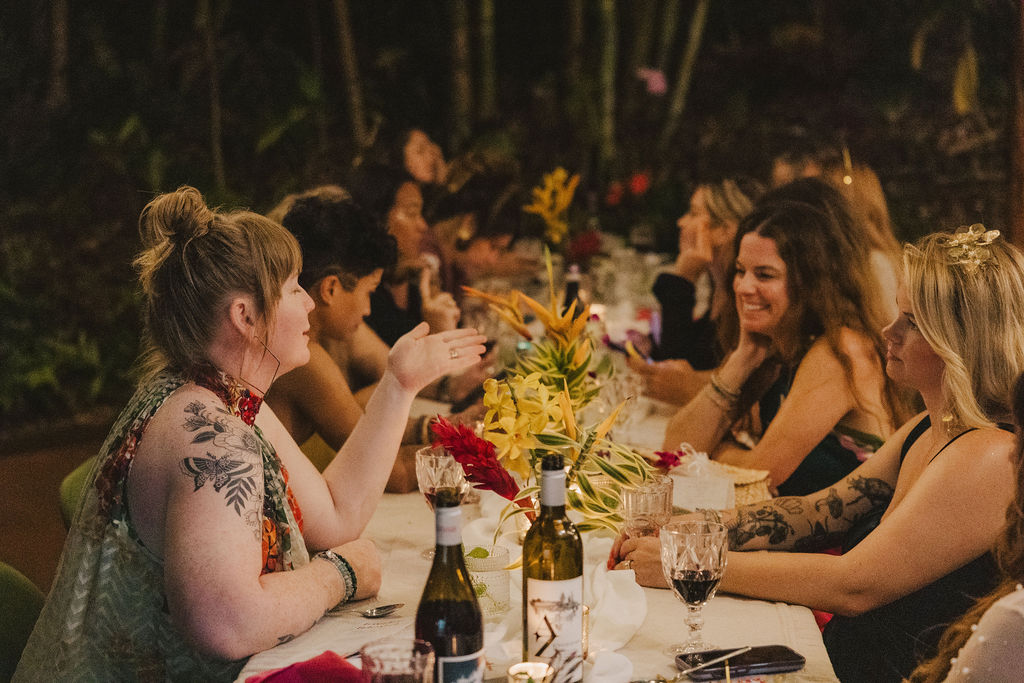 A group of people sit at a decorated outdoor table at night, talking and smiling. The table is adorned with yellow flowers, candles, wine bottles, and glasses, creating a warm and festive atmosphere.