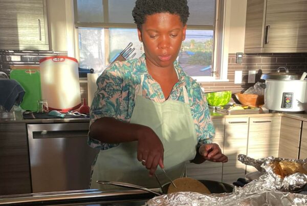 Rethabile Molapo, wearing an apron, prepares food in a kitchen, surrounded by trays of dishes, fresh greens, and ingredients on the counter, with sunlight streaming through a window behind them.