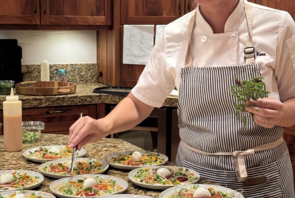 A chef from the lotus chefs team in a striped apron carefully garnishes multiple plated salads in a kitchen with wooden cabinets, smiling as she adds fresh herbs to each dish.