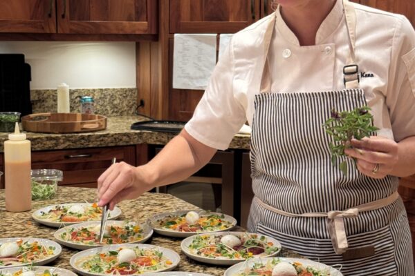 A chef from the lotus chefs team in a striped apron carefully garnishes multiple plated salads in a kitchen with wooden cabinets, smiling as she adds fresh herbs to each dish.