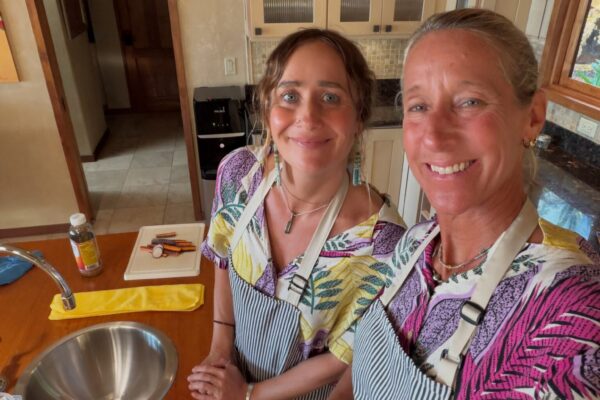 Two people in striped aprons and colorful shirts smile in a kitchen, standing near a counter with fresh fruits and vegetables, ready for meal prep.
