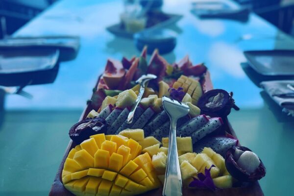 A wooden platter of sliced tropical fruits including mango, dragon fruit, and others sits on a glass table, artfully arranged by Chef Maja Liotta, with a blurred ocean view and dining setup in the background.