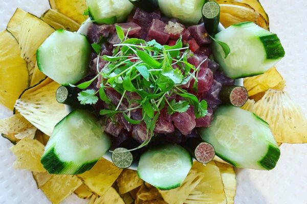 A white plate with sliced cucumber, microgreens, lime halves, diced raw fish in the center, and yellow plantain chips arranged around the edge—an artful presentation inspired by Chef Maja Liotta.