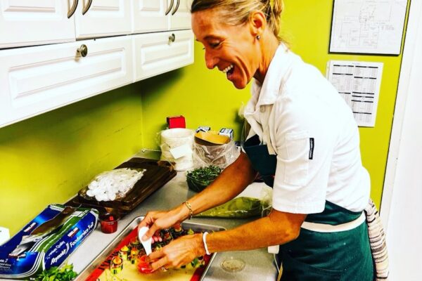 A smiling Chef Maja Liotta in a white uniform and green apron prepares food on a counter in a kitchen with green walls and white cabinets. Various ingredients and utensils are visible around her.