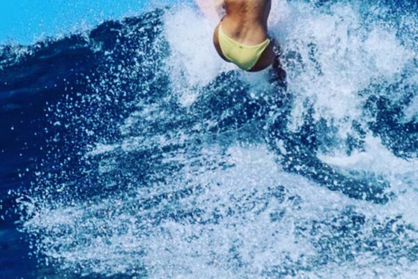 A surfer in a yellow swimsuit rides a wave on a pink surfboard, captured mid-jump against a bright blue sky, with water spraying around them—a scene as vibrant and energetic as one of Chef Maja Liotta’s signature creations.