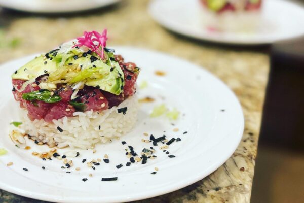 A close-up of a plate with a sushi stack crafted by Chef Maja Liotta, featuring rice, diced raw fish, avocado, sesame seeds, nori, and garnished with a pink edible flower. Other similar plates are blurred in the background.