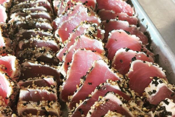 Rows of seared ahi tuna slices coated in sesame seeds are arranged neatly on a metal tray, showing the pink interior and a crust of black and white sesame seeds—a beautifully composed dish crafted by Chef Maja Liotta.