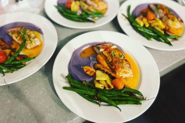 Four white plates with grilled fish, green beans, yellow squash, cherry tomatoes, and a swirl of purple and orange sauces arranged neatly on a countertop showcase Chef Maja Liotta’s exquisite attention to detail.