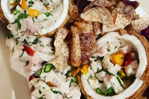Diced fish ceviche, expertly crafted by Chef Maja Liotta, is served in coconut shells with herbs and peppers, accompanied by assorted root vegetable chips on a white plate.