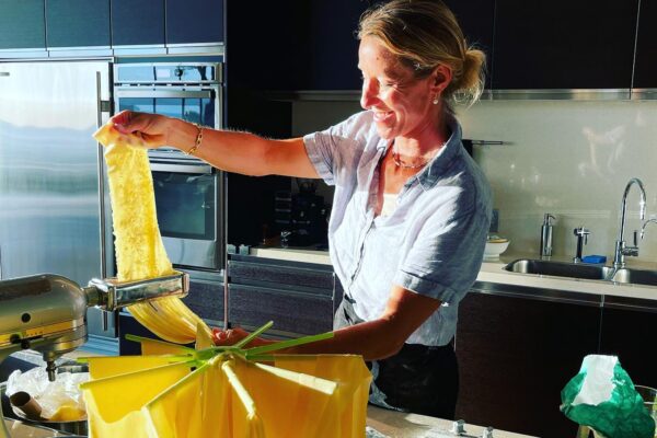 A woman from the Lotus Chefs team smiles while making fresh pasta in a modern kitchen, using a pasta machine. Long pasta sheets hang to dry on a rack nearby under bright sunlight.