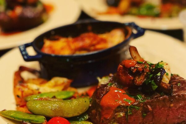 A plated gourmet meal by Chef Maja Liotta features a steak topped with onions and sauce, served alongside snap peas, a cherry tomato, potatoes, and a small casserole dish, with similar plates blurred in the background.