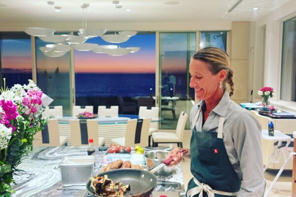 Chef Maja Liotta, wearing an apron, cooks at a kitchen island, smiling while stir-frying vegetables. The modern kitchen has flowers on the counter, with a sunset and ocean visible through large windows in the background.