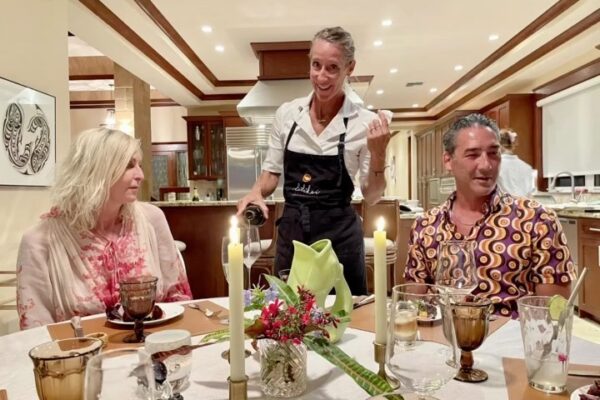 Three people sit at a dining table set with candles and flowers. Chef Maja Liotta, wearing an apron, stands in the middle serving food, while the other two, a man and woman, sit and appear to be conversing with her in a well-lit kitchen.
