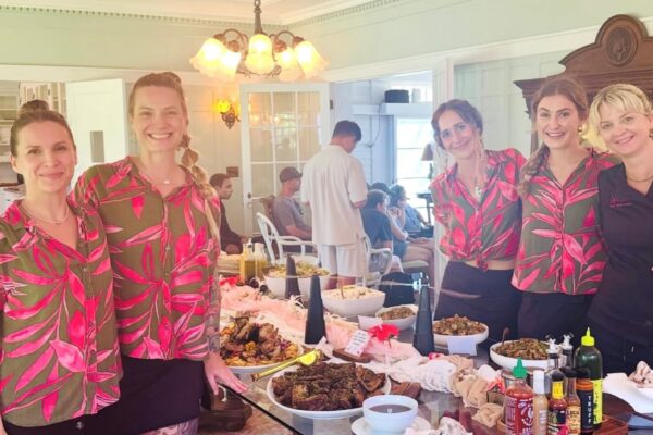 Five women stand smiling by a buffet table with assorted food and condiments in a bright, elegant room. Four wear matching floral shirts and black skirts; one wears black. People are seated in the background.