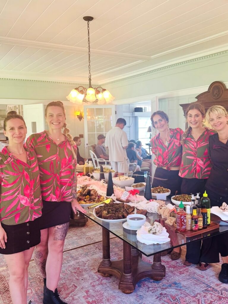 Five women stand smiling by a buffet table with assorted food and condiments in a bright, elegant room. Four wear matching floral shirts and black skirts; one wears black. People are seated in the background.