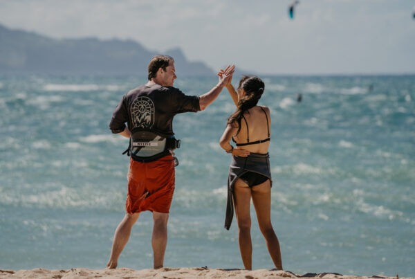A man and woman in swimwear stand on a sandy beach facing the ocean, appearing to high-five. The sky is clear, and there are people kitesurfing in the background—a perfect setting for a corporate retreat catering Maui experience or even an exclusive gathering with a private chef after a day of fun in the sun.