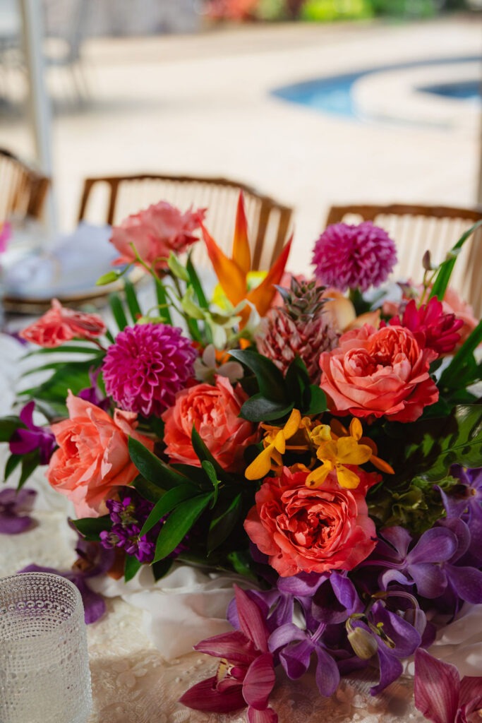 A vibrant floral arrangement with pink roses, orange and yellow flowers, purple orchids, and lush green leaves sits on a table with wooden chairs—perfect for a Maui brunch—with a blurred poolside background.