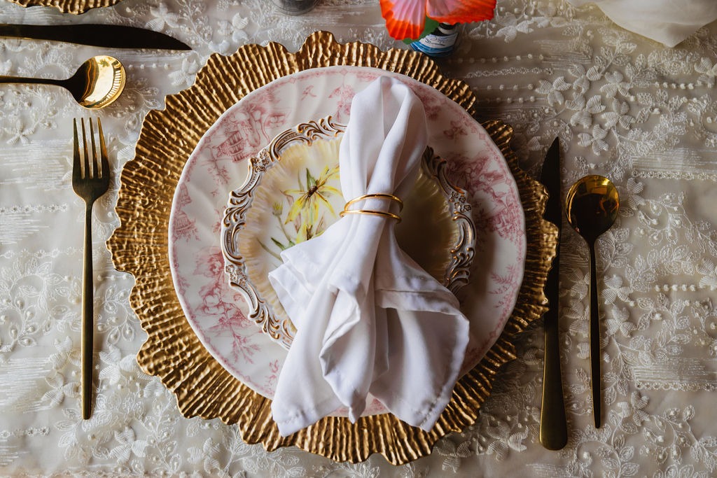 Elegant table setting with a gold-rimmed charger, ornate pink and white plate, white napkin in a gold ring, and gold utensils on a lace tablecloth—perfect for an intimate private chef fine dining experience.