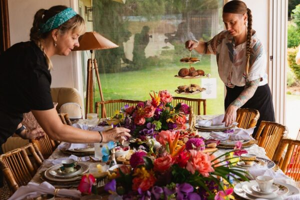 Two women set a dining table decorated with vibrant flowers, elegant china, and tiered trays of pastries in a bright room with large windows, creating an inviting atmosphere reminiscent of a private chef fine dining experience.