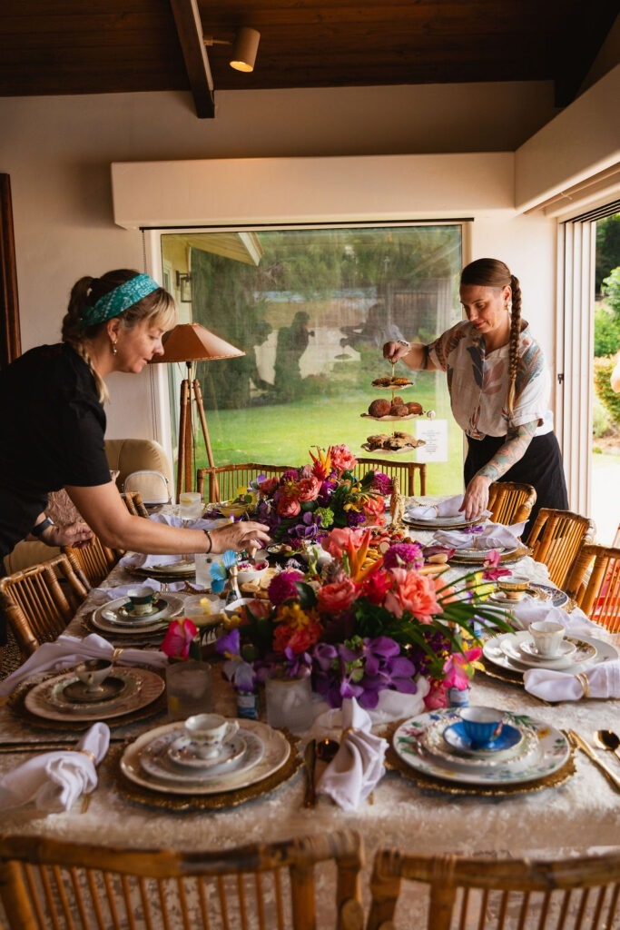Two women set a dining table decorated with vibrant flowers, elegant china, and tiered trays of pastries in a bright room with large windows, creating an inviting atmosphere reminiscent of a private chef fine dining experience.