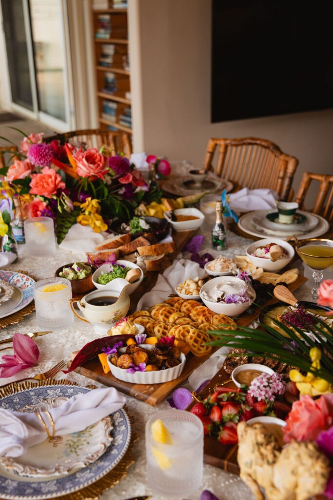 A decorated dining table with vibrant flowers, elegant plates, glasses with lemon water, and assorted foods including breads, vegetables, dips, fruit, and desserts arranged for a festive Maui brunch.
