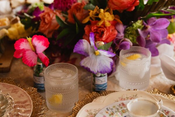 A close-up of an elegant table setting with floral china, gold-rimmed plates, textured glasses with lemon, and a vibrant bouquet of colorful flowers as the centerpiece—perfect for an intimate private chef fine dining experience.