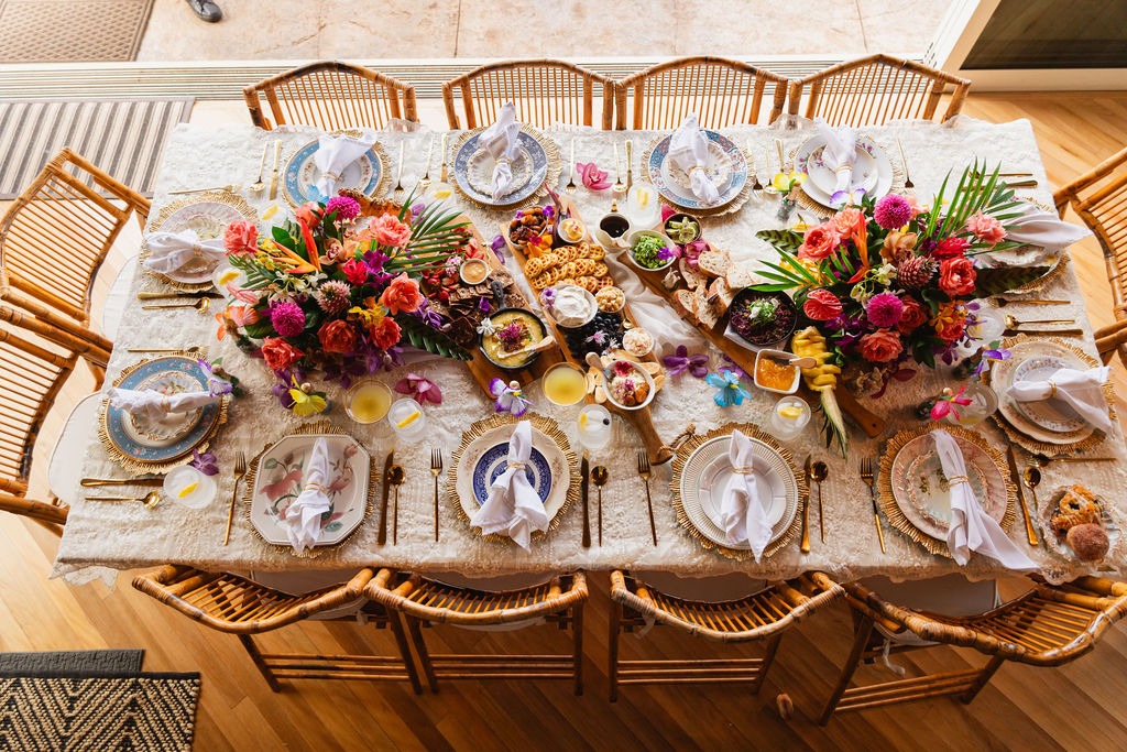 A decorated dining table set for eight with vibrant flower arrangements, ornate plates, gold utensils, napkins folded into shapes, and various food dishes prepared by a private chef viewed from above. Wicker chairs surround the table, creating an atmosphere of fine dining elegance.