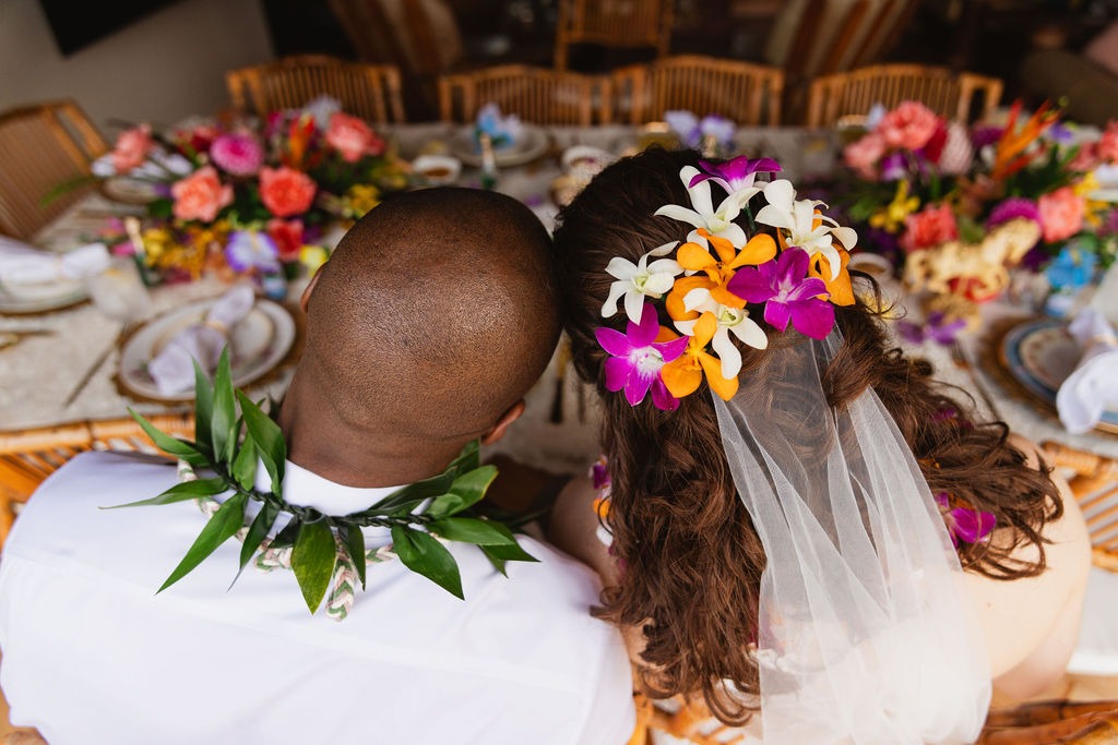 A couple sits together at a decorated table; the man wears a green leaf lei, and the woman has colorful flowers in her hair and a veil. The table is set with vibrant floral arrangements and elegant place settings, creating an enchanting atmosphere perfect for private chef fine dining.
