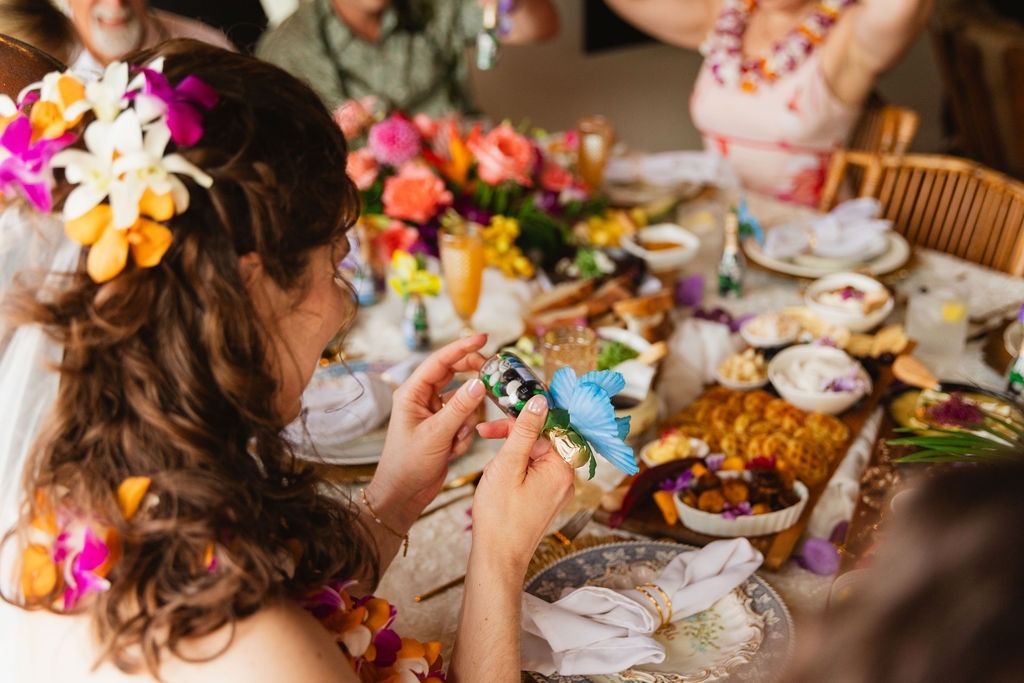A woman with flowers in her hair sits at a decorated table, holding a small gift. The table, arranged for private chef fine dining, is filled with food, drinks, and colorful flowers. Other people are seated nearby, partially visible in the background.