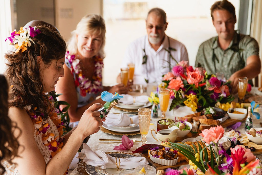 A group of people wearing leis sit around a table decorated with colorful flowers, food, and drinks prepared by a private chef. One woman examines a small toy figurine while others smile and enjoy the festive fine dining atmosphere.