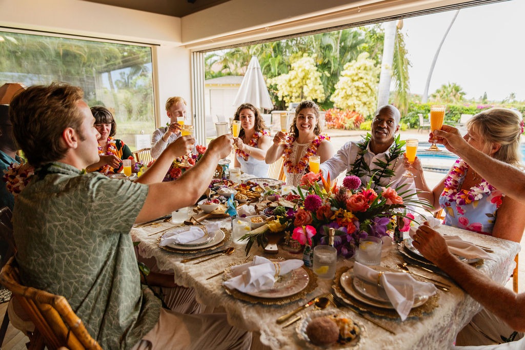 A group of people wearing leis sit around a festive, flower-decorated table, raising glasses in a toast near a sunny window with a pool and palm trees in the background, savoring an exquisite meal crafted by a private chef for an unforgettable fine dining experience.