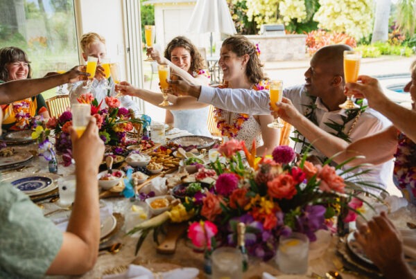 A group of people sit around a table with colorful flowers and food, raising glasses in a celebratory toast, smiling and enjoying a festive meal together indoors by a sunny window—a memorable experience of fine dining curated by a private chef.