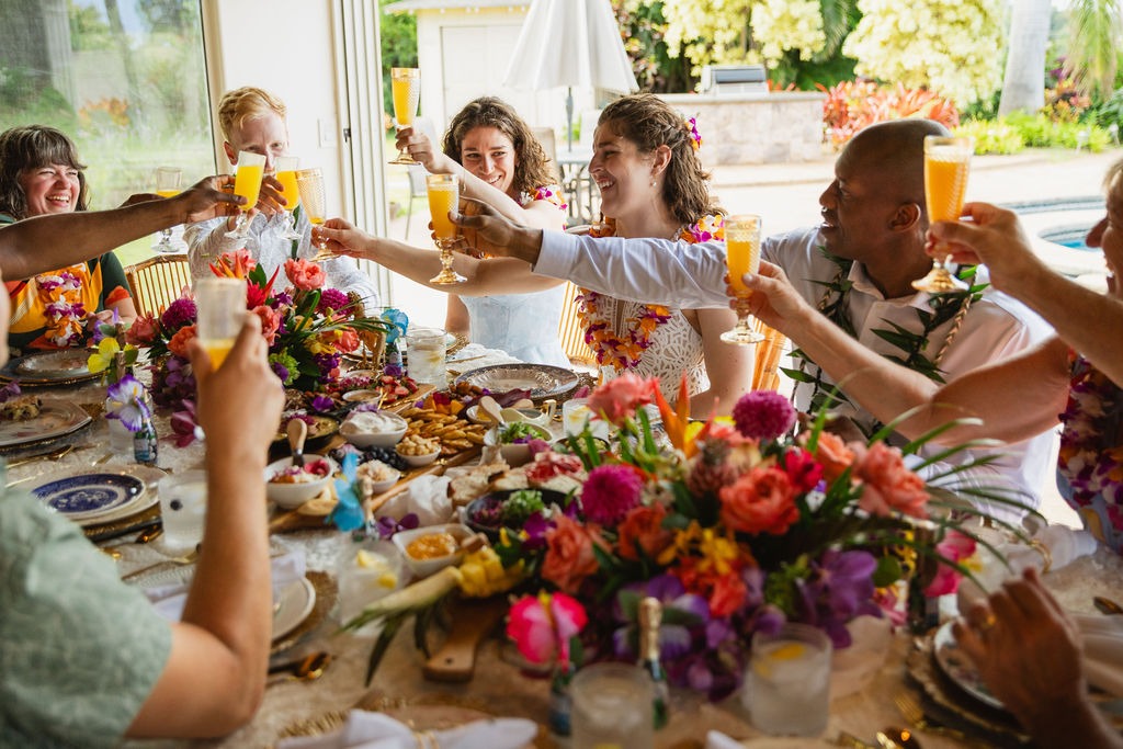 A group of people sit around a table with colorful flowers and food, raising glasses in a celebratory toast, smiling and enjoying a festive meal together indoors by a sunny window—a memorable experience of fine dining curated by a private chef.