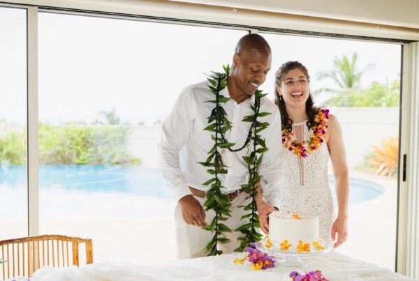 A smiling couple wearing leis stands by a white cake decorated with flowers in front of a window overlooking a pool and greenery, celebrating their special day with the personalized touch of an elopement chef.