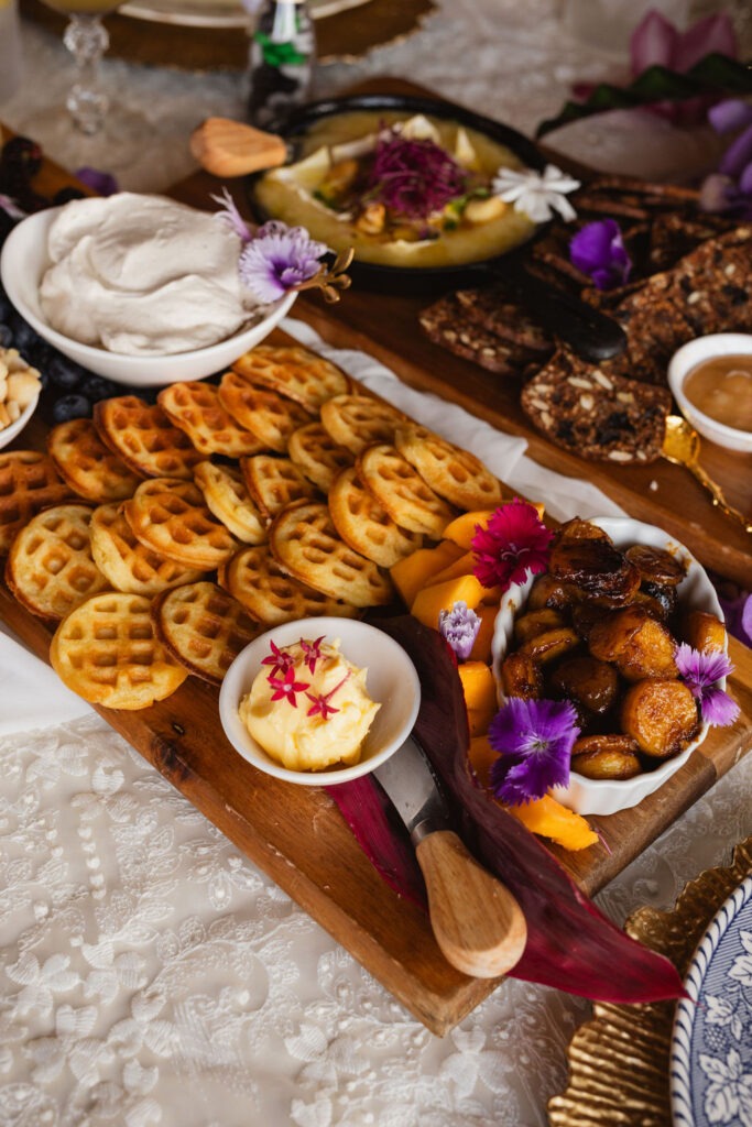 A wooden platter featuring mini waffles, whipped cream, butter, caramelized bananas, fresh peaches, and edible flowers evokes the vibrant flavors of a Maui brunch. Set on a lace tablecloth with other dishes partially visible, this delightful spread brings a touch of island elegance to your table.