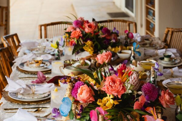 A dining table set for a formal meal, decorated with vibrant floral centerpieces, gold-rimmed plates, glasses, folded napkins, and bamboo chairs in a sunlit room—perfect for an intimate private chef fine dining experience.