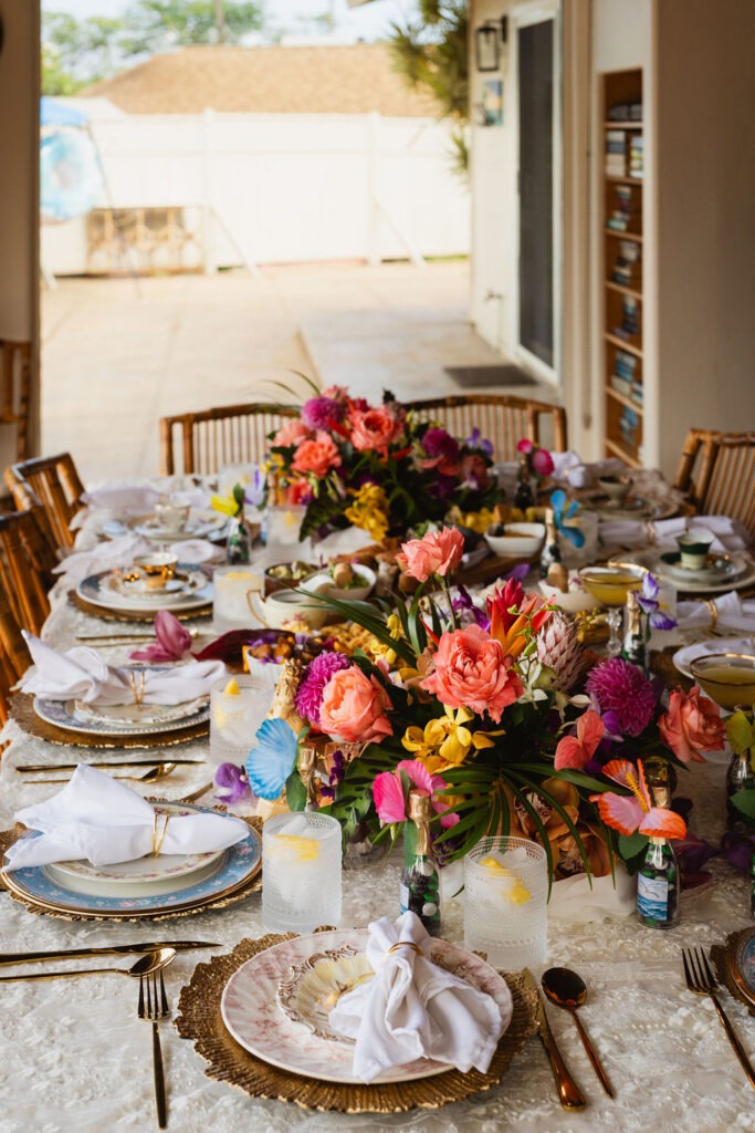 A dining table set for a formal meal, decorated with vibrant floral centerpieces, gold-rimmed plates, glasses, folded napkins, and bamboo chairs in a sunlit room—perfect for an intimate private chef fine dining experience.