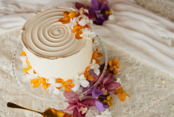 A white frosted cake with a spiral design on top, decorated with orange, white, and purple flowers, sits on a glass stand—perfect for an intimate elopement or microwedding celebration. Flowers are also scattered on the tablecloth nearby, adding to the elegant display. A gold cake server is visible, reflecting the refined touch of professional chef services.
