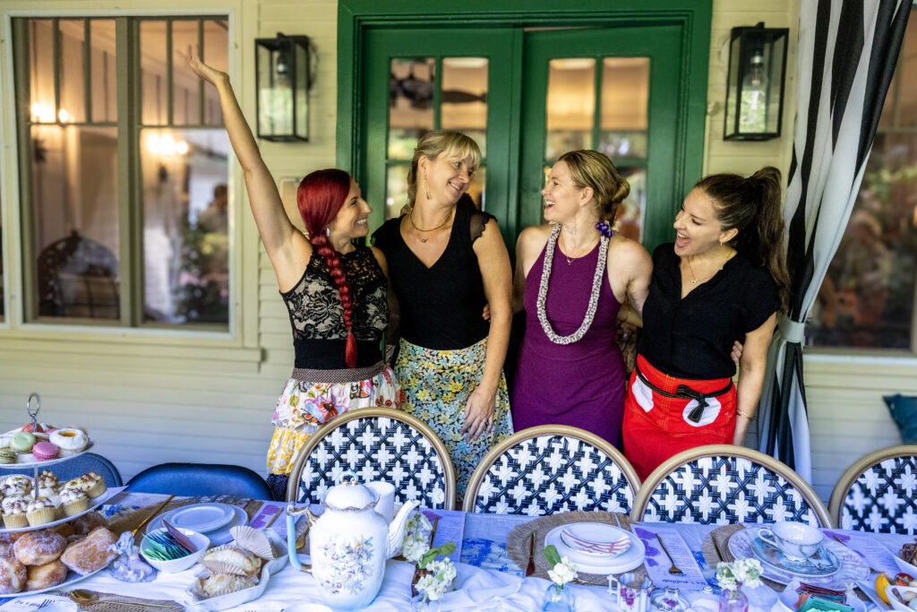 Four women stand and laugh together behind a table set with tea, pastries, and desserts on a porch with green French doors and patterned chairs, enjoying a cheerful tea party—a moment beautifully captured by Maui Photographers.