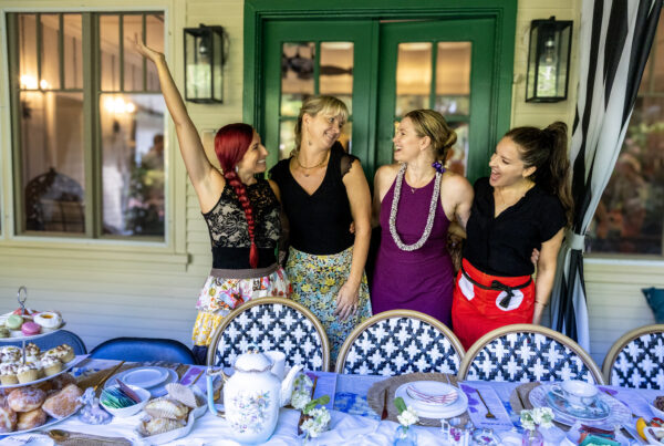 Four women stand and laugh together behind a table set with tea, pastries, and desserts on a porch with green French doors and patterned chairs, enjoying a cheerful tea party—a moment beautifully captured by Maui Photographers.